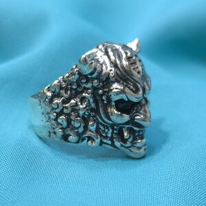 Könnte beinhalten: Ein silberner Ring mit einem detaillierten Design eines Dämonengesichts. Der Ring liegt auf einem blauen Hintergrund.