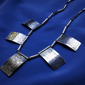以下が含まれることがあります： 複雑なエンボス加工が施された5つの長方形のペンダントが付いたシルバーネックレス。ネックレスは青い背景の上に置かれています。