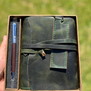 May include: A green leather journal with a pen in a box. The journal has a green leather strap and a bookmark. The pen is silver and brown.