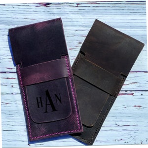 May include: Two leather pencil cases, one purple and one brown, with a monogrammed initial "A" on the purple case. The cases have a flap closure and are stitched with pink thread.