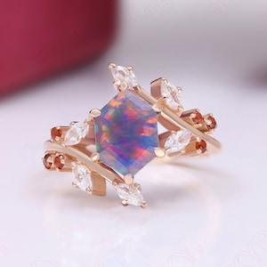 Puede incluir: Un anillo de compromiso de oro rosa con una piedra de ópalo hexagonal rodeada de piedras preciosas más pequeñas de color claro y naranja. El anillo presenta un diseño delicado en forma de hoja.