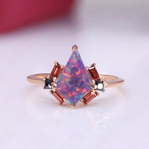 May include: A rose gold engagement ring with a large, iridescent, purple and pink opal gemstone in a geometric setting. The ring is accented with smaller black and red gemstones.
