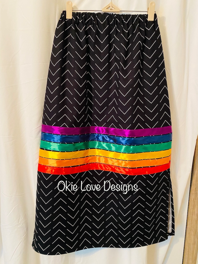 May include: Black skirt with a white chevron pattern. The skirt features a horizontal band of rainbow-colored ribbons. The text "Okie Love Designs" is printed on the skirt.