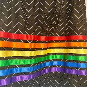 May include: A black fabric with a white chevron pattern. The bottom of the fabric is adorned with horizontal ribbons in a rainbow of colors: red, orange, yellow, green, blue, and purple.