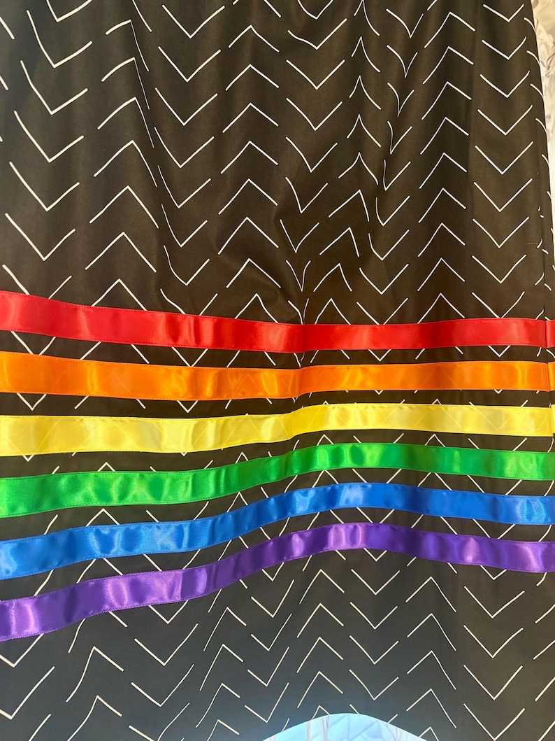 May include: A black fabric with a white chevron pattern. The bottom of the fabric is adorned with horizontal ribbons in a rainbow of colors: red, orange, yellow, green, blue, and purple.