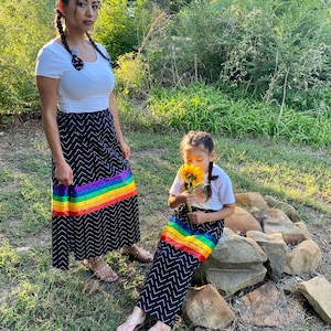 May include: Two people wearing matching black skirts with a white chevron pattern and a rainbow stripe. They are also wearing white tops and sandals. One person is holding a sunflower. The setting is outdoors with green foliage.