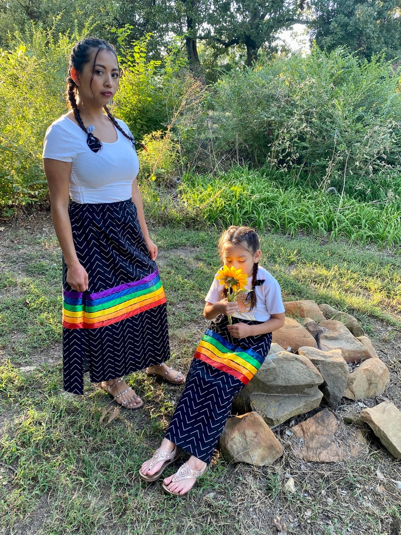 May include: Two people wearing matching black skirts with a white chevron pattern and a rainbow stripe. They are also wearing white tops and sandals. One person is holding a sunflower. The setting is outdoors with green foliage.