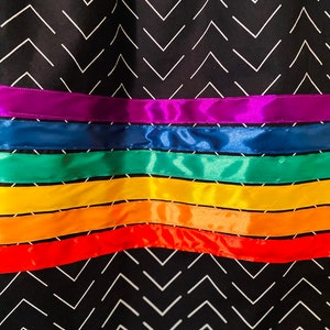 May include: Close-up of a fabric with a black chevron pattern and horizontal rainbow-colored ribbons. The ribbons are in shades of red, orange, yellow, green, blue, and purple. The fabric has a repeating white chevron design.