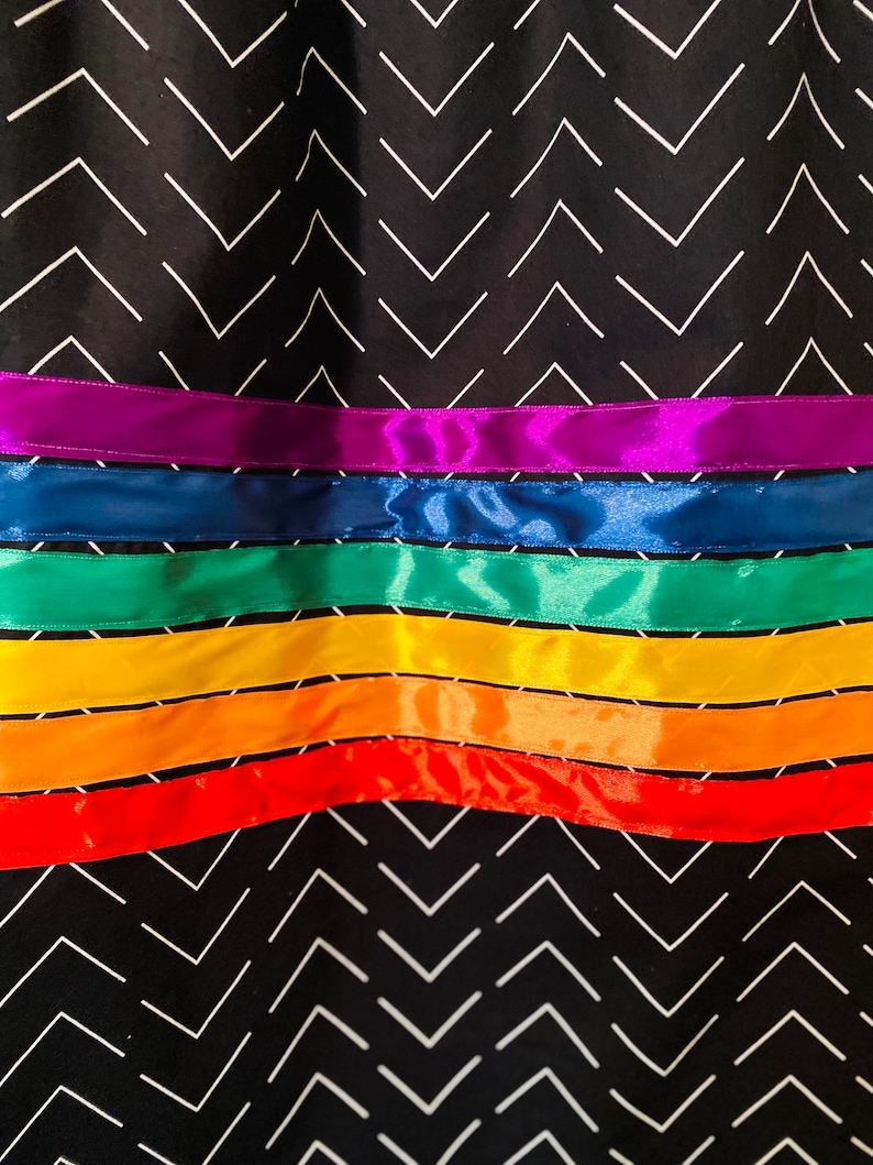 May include: Close-up of a fabric with a black chevron pattern and horizontal rainbow-colored ribbons. The ribbons are in shades of red, orange, yellow, green, blue, and purple. The fabric has a repeating white chevron design.