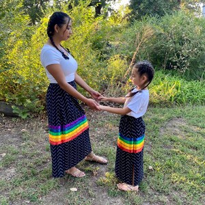 May include: Two people wearing matching navy blue skirts with a white chevron pattern and a rainbow stripe at the hem. They are also wearing white tops. The skirts are long and flowing, and the rainbow stripe adds a pop of color.