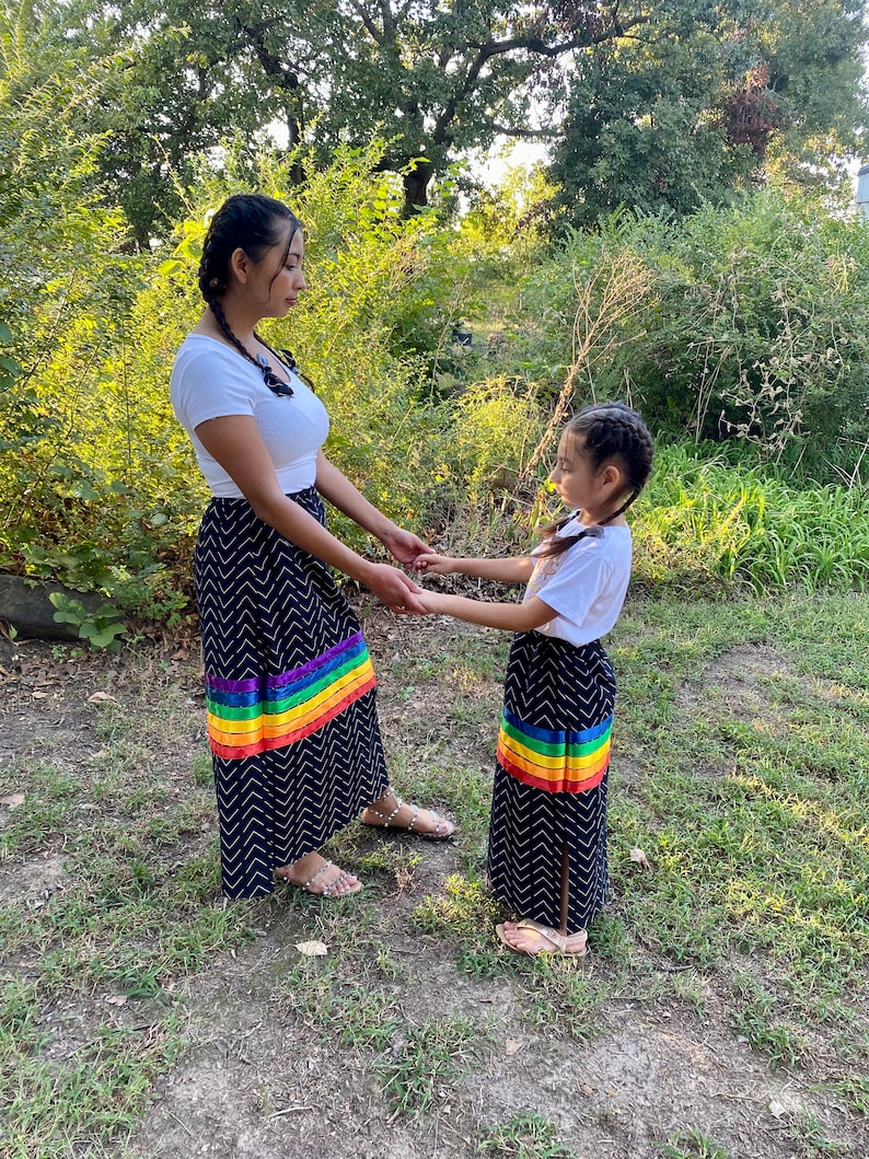 May include: Two people wearing matching navy blue skirts with a white chevron pattern and a rainbow stripe at the hem. They are also wearing white tops. The skirts are long and flowing, and the rainbow stripe adds a pop of color.