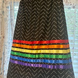 May include: A black skirt with a white chevron pattern. The skirt features horizontal bands of rainbow-colored ribbon. The skirt is hanging on a black hanger in front of a window with sheer curtains.