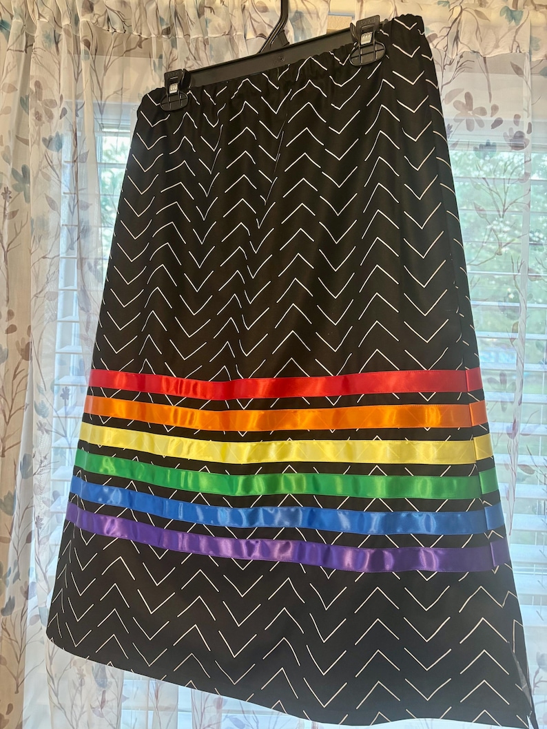 May include: A black skirt with a white chevron pattern. The skirt features horizontal bands of rainbow-colored ribbon. The skirt is hanging on a black hanger in front of a window with sheer curtains.