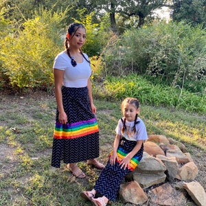 May include: Two people wearing matching black skirts with a chevron pattern and a rainbow stripe. They are also wearing white tops and sandals. The photo is taken outdoors with trees and bushes in the background.