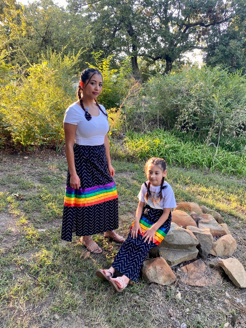 May include: Two people wearing matching black skirts with a chevron pattern and a rainbow stripe. They are also wearing white tops and sandals. The photo is taken outdoors with trees and bushes in the background.