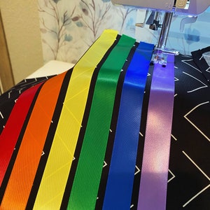 May include: A sewing machine stitching colorful satin ribbons onto a black fabric with a white geometric pattern. The ribbons are arranged in a rainbow spectrum: red, orange, yellow, green, blue, and purple. The machine's needle is in action.