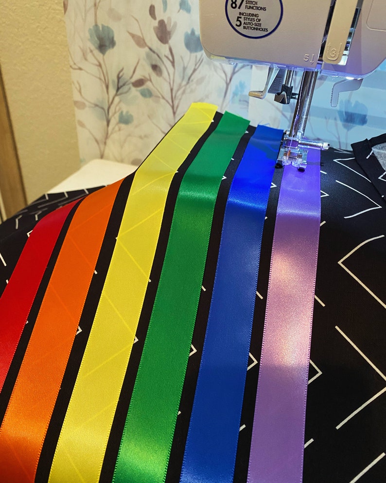 May include: A sewing machine stitching colorful satin ribbons onto a black fabric with a white geometric pattern. The ribbons are arranged in a rainbow spectrum: red, orange, yellow, green, blue, and purple. The machine's needle is in action.