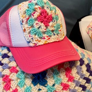 Peut inclure: Casquette de camionneur rose avec un panneau avant blanc et un motif carré crocheté. Le carré crocheté présente une fleur rose au centre, entourée d'accents bleus et verts. La casquette a une visière rose et un dos en filet.