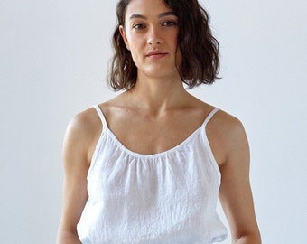 May include: A white linen camisole top with thin straps and a gathered neckline. The top is paired with high-waisted, light blue pants with an elastic waistband. The outfit is simple and casual, suitable for warm weather.