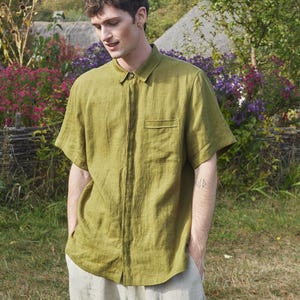 May include: A green linen shirt with a single chest pocket and short sleeves. The shirt is buttoned up and the model is standing with their hands in their pockets.