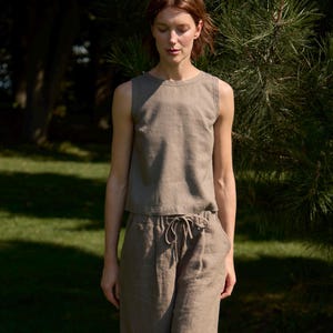 Op de afbeelding: Een tweedelige linnen outfit in een gedempte olijfgroene kleur. De mouwloze top en de broek met wijde pijpen hebben een relaxte pasvorm. De broek heeft een taille met trekkoord. De outfit wordt gedragen met neutrale sandalen. Het model staat in het gras.