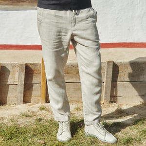 May include: A person wearing a black shirt and light grey linen trousers. The trousers have a drawstring waist and are cuffed at the ankles. The person is wearing white trainers.