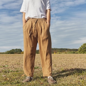 May include: A pair of brown linen trousers with a drawstring waist and a relaxed fit. The trousers are being worn by a person standing in a field.