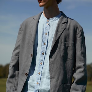 May include: A grey linen blazer with two brown buttons, worn over a light blue button-down shirt. The outfit is completed with matching grey trousers. The ensemble is suitable for a casual or semi-formal setting.