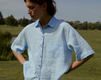 May include: A light blue linen shirt with short sleeves and a front pocket. The shirt has wooden buttons and is paired with beige linen pants. The model is standing on a green grassy field with a pond and trees in the background.