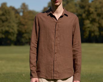 Può includere: Una camicia marrone a maniche lunghe con bottoni, abbinata a pantaloni beige. La camicia ha un colletto classico ed è realizzata in un tessuto strutturato. I pantaloni sono a gamba larga e di colore chiaro. Lo sfondo è un campo verde e alberi.