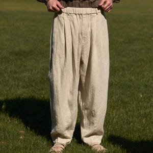 Könnte beinhalten: Eine Person trägt einen braunen Kapuzenpullover und eine beige Leinenhose. Die Hose hat eine lockere Passform mit Falten an der Taille. Das Outfit wird mit Sandalen auf einer Wiese getragen.