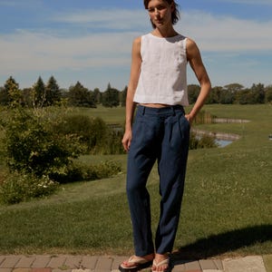 May include: A woman wearing a white sleeveless top and navy blue pants. The pants have a pleated front. She is wearing flip-flops and standing on a brick walkway. The background is a green field and blue sky.
