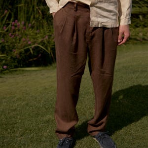 May include: A person wearing a light beige button-down shirt and dark brown pleated trousers. The person is also wearing dark blue boat shoes with a woven sole. The outfit is set against a backdrop of green grass and foliage.