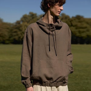 Könnte beinhalten: Ein braunes Kapuzensweatshirt mit Kordelzug am Ausschnitt und elastischen Bündchen und Saum. Das Sweatshirt wird mit beigefarbenen Leinenhosen kombiniert. Das Model steht auf einer grünen Wiese mit blauem Himmel im Hintergrund.