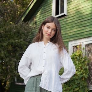 May include: A young person wearing a white linen shirt and green linen pants stands in front of a green wooden building with white trim. The building has a window with white trim and a door with white trim.