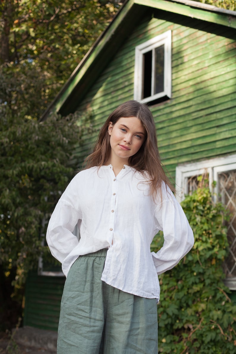 May include: A young person wearing a white linen shirt and green linen pants stands in front of a green wooden building with white trim. The building has a window with white trim and a door with white trim.