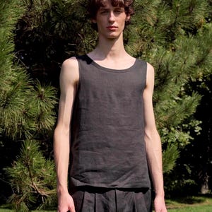 May include: A dark grey vest top and matching pleated trousers. The sleeveless top has a rounded neckline. The outfit is made of a soft, textured fabric. The model is standing in front of a green, leafy background.