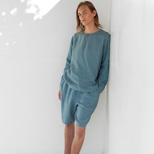 May include: A person wearing a light blue linen shirt and matching shorts. They are standing against a white wall with their arms at their sides. They are wearing brown leather shoes.