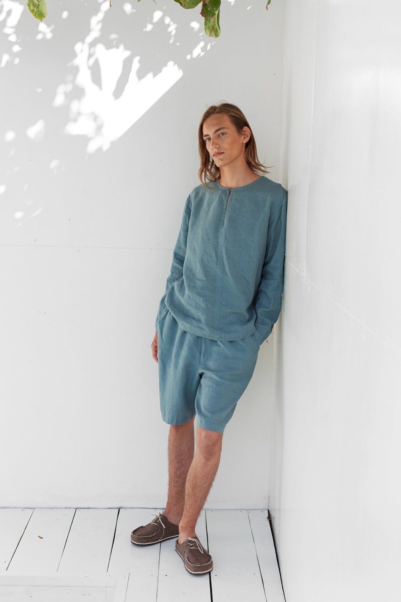May include: A person wearing a light blue linen shirt and matching shorts. They are standing against a white wall with their arms at their sides. They are wearing brown leather shoes.