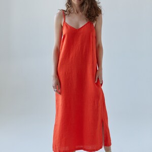 May include: A woman wearing a bright orange maxi dress with thin straps. The dress has a simple, straight cut and a slight slit in the front. She is wearing light green sandals.