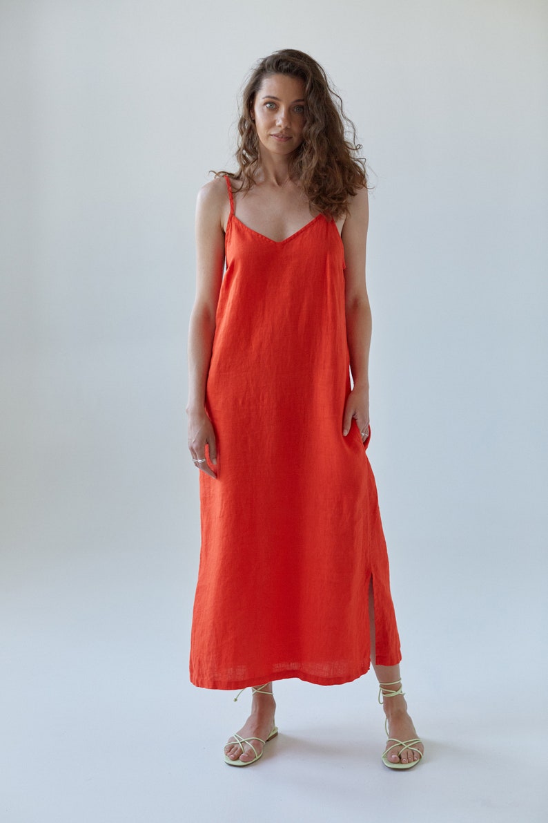 May include: A woman wearing a bright orange maxi dress with thin straps. The dress has a simple, straight cut and a slight slit in the front. She is wearing light green sandals.