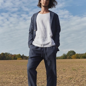 May include: A man wearing a dark blue linen blazer and matching pants. He is standing in a field with a cloudy sky in the background.
