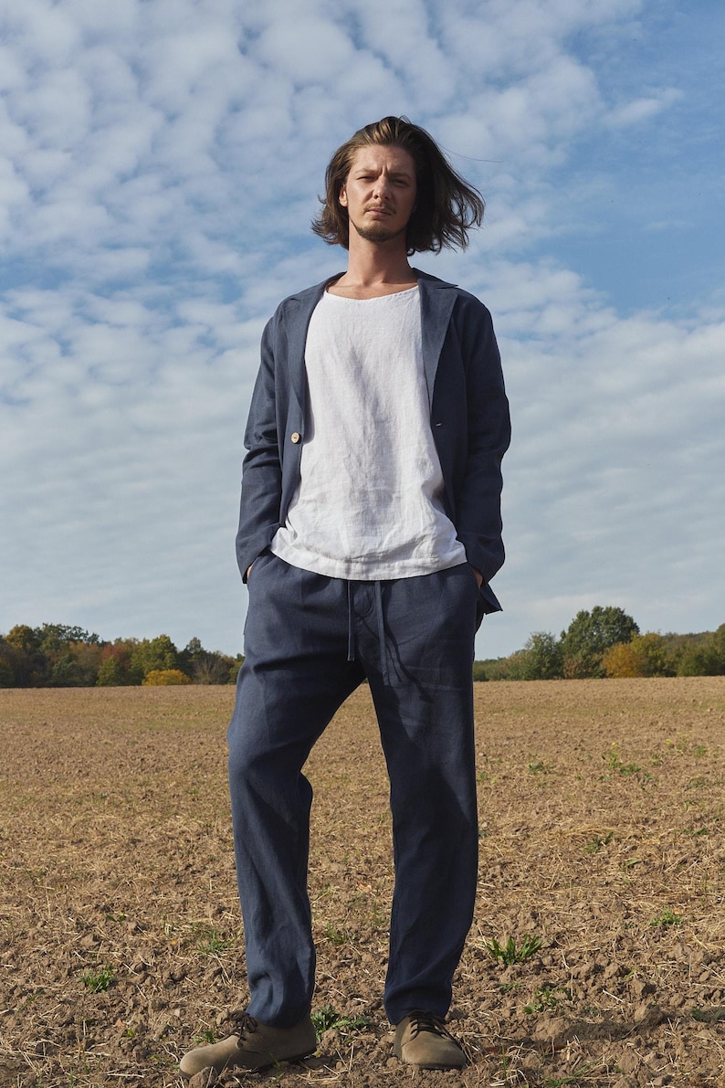May include: A man wearing a dark blue linen blazer and matching pants. He is standing in a field with a cloudy sky in the background.