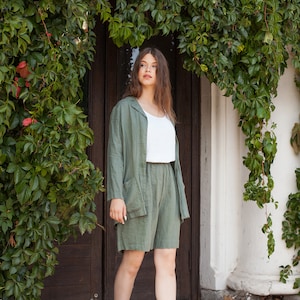 May include: A woman wearing a green linen shorts set with a white tank top and black combat boots. She is standing in front of a wooden door with ivy growing over it.
