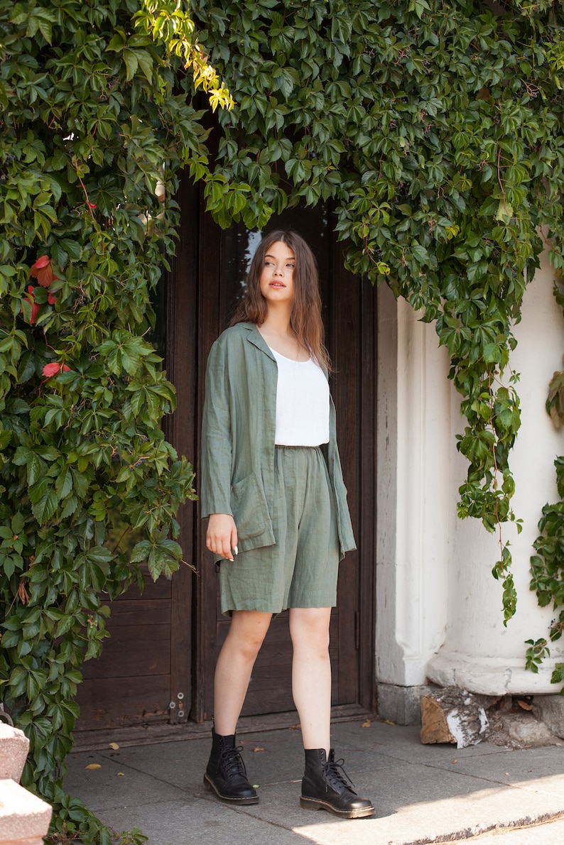 May include: A woman wearing a green linen shorts set with a white tank top and black combat boots. She is standing in front of a wooden door with ivy growing over it.