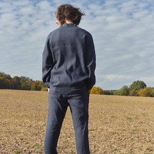 May include: A person wearing a dark blue linen shirt and matching pants standing in a field. The person is facing away from the camera and looking at the horizon.