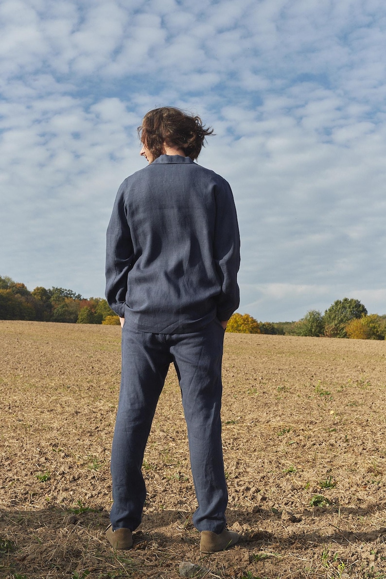 May include: A person wearing a dark blue linen shirt and matching pants standing in a field. The person is facing away from the camera and looking at the horizon.