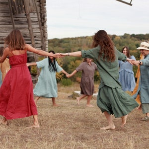 May include: A group of people wearing long, flowing dresses in various colours, including red, blue, green, and yellow, are holding hands and dancing in a circle in a field. They are smiling and laughing as they dance. There is a wooden building in the background.