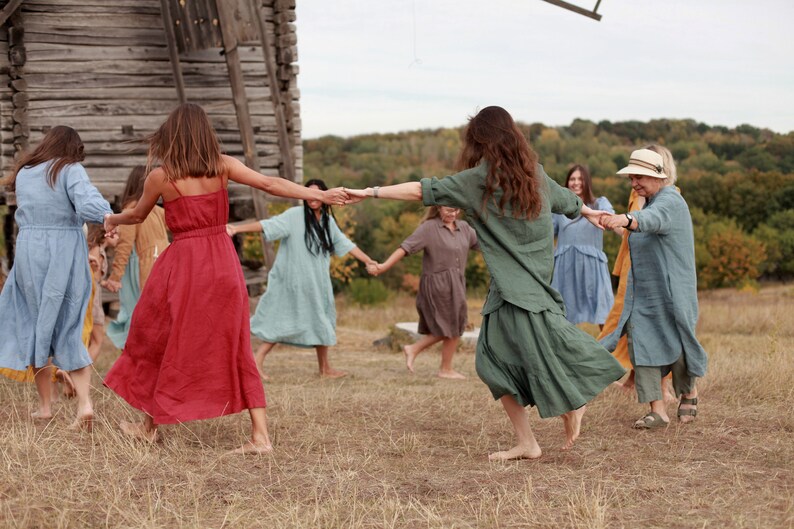 May include: A group of people wearing long, flowing dresses in various colours, including red, blue, green, and yellow, are holding hands and dancing in a circle in a field. They are smiling and laughing as they dance. There is a wooden building in the background.