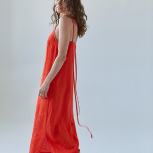May include: A woman wearing a bright orange maxi dress with thin straps. The dress has a side slit and a long tie that hangs down the back. She is wearing light green sandals.
